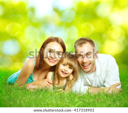 Happy smiling young family father, mother and little daughter having fun outdoors, playing together in summer park. Mom, Dad and kid laughing, lying on green grass, enjoying nature outside. Sunny day
