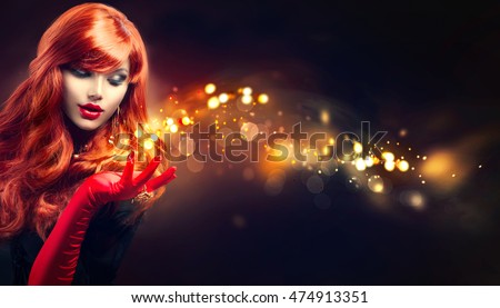 Beauty Glamour Woman with golden sparks magic in her hand. Long red wavy hair. Fashion Lady with Beautiful Luxury Hairstyle, makeup, accessories. Isolated on black. Retro Red Silk Gloves, dark dress