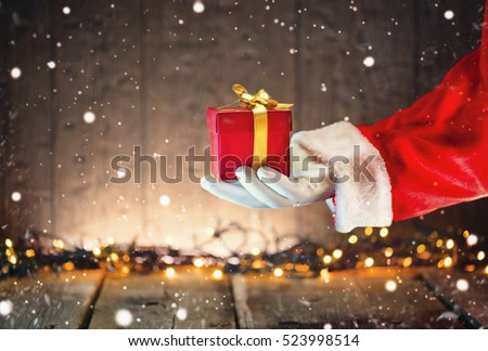 Santa Claus hand holding Christmas Gift box over wooden background. Proposing product. Advertisement gesture presenting point. Decorated Christmas tree with Winter Holiday Gifts.