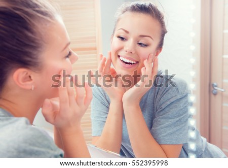 Beauty skin care. Young beautiful teenage girl touching her face before the mirror, enjoying her clean skin. Pretty woman touching her cheek and smiling. Perfect pure skin. Fresh Clean Skin. Skincare