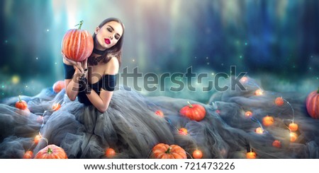 Halloween Witch with a carved Pumpkin and magic lights in a dark forest. Beautiful young surprised woman in witches hat and costume holding pumpkin. Wide Halloween party art design