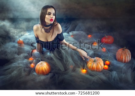 Halloween Witch with Pumpkins and magic lights in a dark room. Beautiful young sexy woman in witches costume sitting with pumpkins and candles. Witchcraft scene art design Halloween party art design.