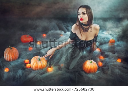 Halloween Witch with Pumpkins and magic lights in a dark room. Beautiful young sexy woman in witches costume sitting with pumpkins and candles. Witchcraft scene art design Halloween party art design.