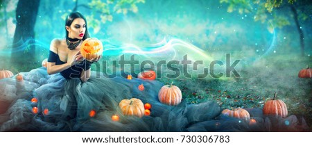 Halloween Witch with a carved Pumpkin and magic lights in a dark forest. Beautiful young surprised woman in witches costume holding pumpkin. Wide Halloween party wide angle art design