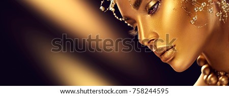 Fashion art Golden skin Woman face portrait closeup. Gold jewellery, jewelry and accessories. Beauty gold eyes, Lips and Skin and hair. Model girl with holiday golden Glamour shiny professional makeup