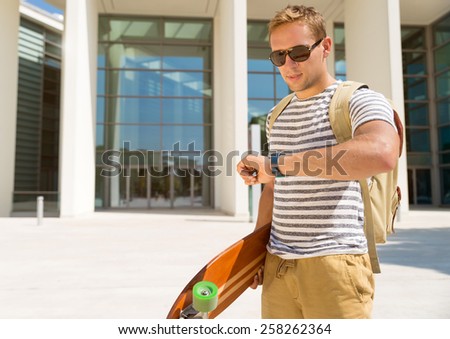 Handsome hipster guy looking at his watch for the next date. Checking time for meeting with luggage and longboard on city background