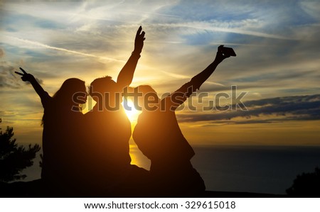 Silhouettes of young group of people sitting during sunset and take a selfie with smartphone. Hipster best friends having fun on low cost road trip. Concept of freedom and technology around the world.