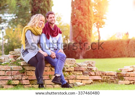 Young couple in love on dating and share good time together. Happy girlfriend sitting with his boyfriend on bricks wall and holding hands. Magic moments in a sunny autumn afternoon color tones