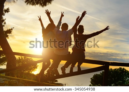 Rear view of three couple best friends travelers put hands up at sunset. Young  relaxing hipster wanderers enjoying exclusive alternative destination. Holiday life moment at warm orange light