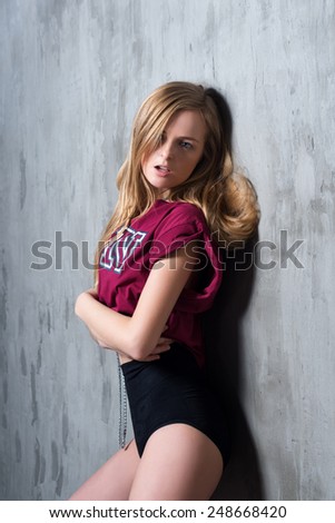 Sexy young blonde woman in black luxury lingerie and sports t-shirt posing against grungy gray wall. Passion and desire pose. Attractive woman with long precious chain