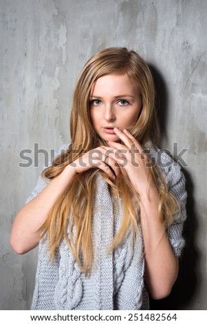 Sexy young longhair blonde woman in knitted sweater posing against grungy gray wall. Scared or resentful attractive girl hiding behind a lock of her hair.