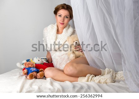 Young beautiful fashion pregnant woman sitting on the bed with toys for her baby on white background.