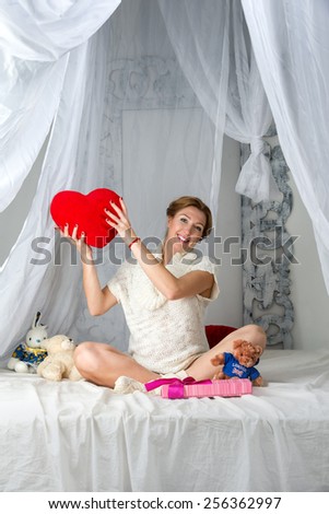 Young beautiful fashion pregnant woman sitting on the bed with toys for her baby on white background.