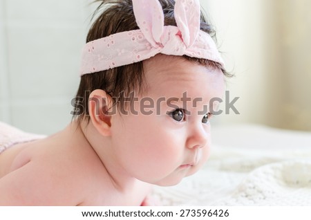 Portrait of cute sweet little newborn baby girl in pink head bandage bow looking with big beautiful hazel brown eyes, curious glance, photographed side-face. The depth of field is short.