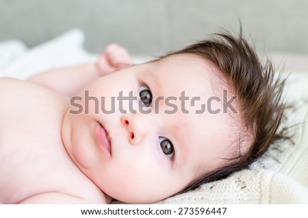 Portrait of cute sweet little newborn baby girl looking at camera with big beautiful hazel brown eyes, lying on white woolen blanket on a bed at home