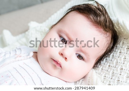 Portrait of cute sweet little newborn baby girl with black brunette hair looking with big beautiful hazel brown eyes and lying on white woolen blanket on a bed at home