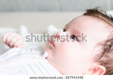 Portrait of cute sweet little newborn baby girl in side-face with black hair looking with big beautiful hazel brown eyes, lying on white woolen blanket on a bed at home. The depth of field is short.