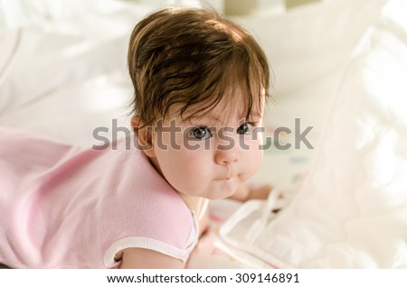Cute sweet little baby girl with black brunette hair in nice pink romper suit lying on her bed at home and looking at the camera with smart and curious beautiful hazel brown eyes