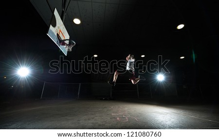 Basketball court at night with lights on, basketball player jumping and aiming at hoop