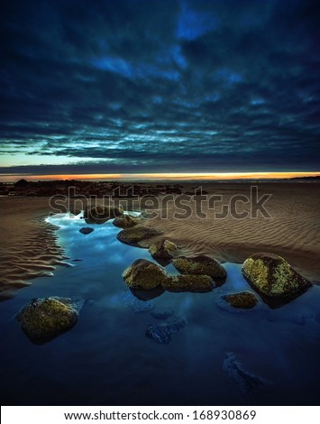 Very Calm And Tranquil Beach Scene With Rocks In A Pure Blue Water Puddle With Sand Leading Out To The Horizon, With Dark Blue Clouds And A Red Sunset.