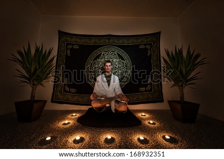 A man sitting on a zafu cushion with in a white robe with his eyes closed doing a  meditation ritual. There are plants, candles, and a tapestry behind him on the wall.
