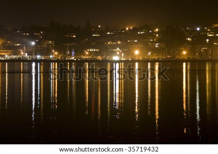 A harbor/bay with lots of boats and lit up houses at night with a reflection of them on the water below.