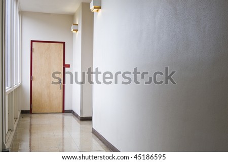 An empty hallway leading to a closed door. Windows let light into the hall coming from the left.