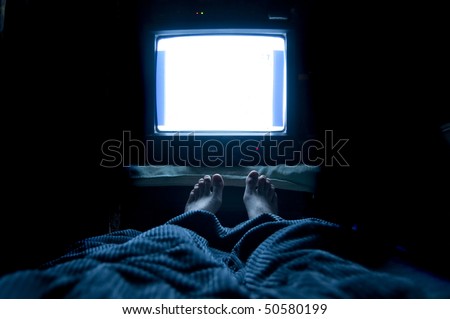 Person watches TV at night in his bed with his feet sticking up out of the blankets. The TV screen is blank white