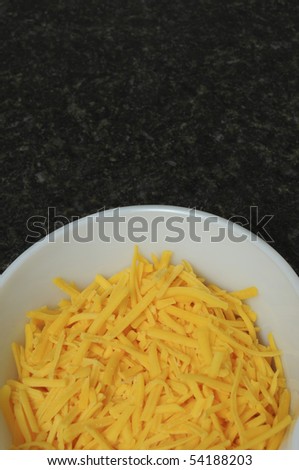 An aerial view of a white bowl containing yellow cheddar cheese on a black counter top
