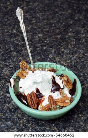 A sundae with pecans, hot chocolate fudge, whip cream, and ice cream inside of a green dessert bowl with a long spoon sitting on a kitchen counter top.