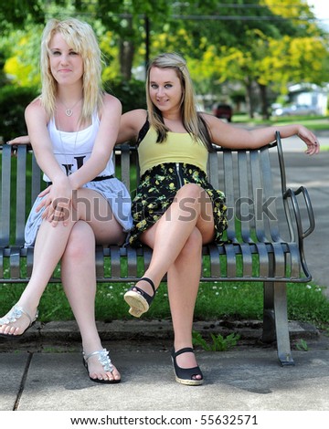 Two girl friends sitting on a metal bench, smiling at the camera. They can be seen as being flirtatious, happy or welcoming.