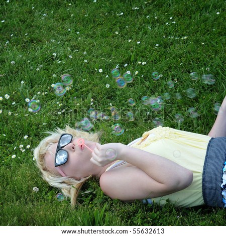 Young white Caucasian teenage girl laying in grass blowing bubbles with her sunglasses on, looking up at the sky.