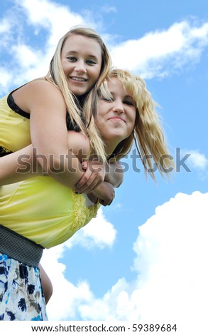 Two beautiful and attractive happy blond teenage girls giving a piggyback ride in the sunshine with clouds and sky in the background.