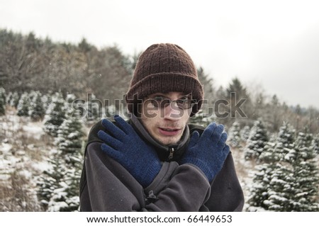 Young man shivers rubbing his hands together in the snow. He is wearing a beanie and a sweater.