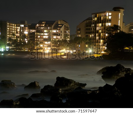 Beautiful ocean sea rocks with misty fog and an illuminated ocean-view hotel resort off in the distance on the horizon.