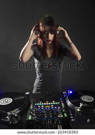 Pretty Young Female Disc Jockey in Casual Gray Shirt Standing at her Deck While Listening to Music using Headphone, Isolated on Gray Background.
