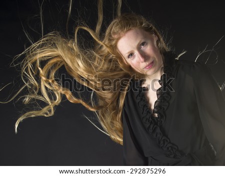Portrait of Smiling Woman Wearing Frilly Black Dress with Long Blond Hair Blowing in Strong Wind, on Black Background