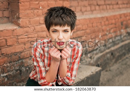 Looking into the camera. Young woman plaid shirt jeans near brick wall.
