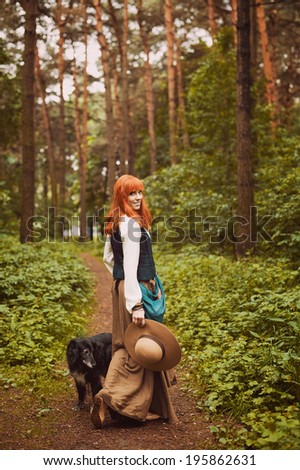 Redhead hippie walks in summer forest and playing with the dog. Young woman with long hair in a long skirt and blouse in Boho style. Photo of romantic girl in national dress.