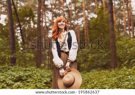 Redhead hippie walks in summer forest. Young woman with long hair in a long skirt and blouse in Boho style. Photo of romantic girl in national dress.