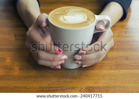 A female\'s hands with heart nail art is holding a cafe with latte art. The sunshine is slanting through the window
