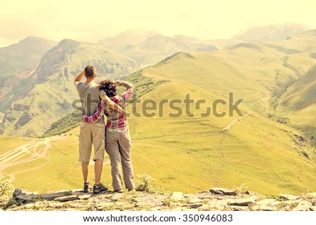Rear view, couple hikers looking into the distance, is standing on the top of mountain. Background is mountains, valley and cloudy sky.