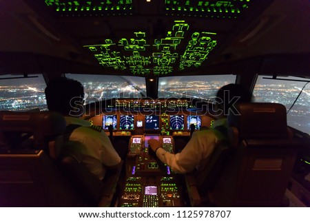 Two pilots are flying the airplane in approach phase to the runway in night time while raining. Cityscape and airport are seen outside cockpit. Pilots and airplane instruments are inside cockpit.