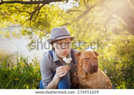 happy young woman in a hat with dog Shar Pei sitting in the field in sunset light and blowing on a dandelion flowers, best friends forever, people concept