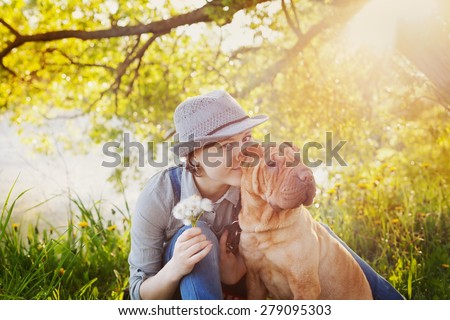 happy young woman in denim overalls and hat with a bouquet of dandelions kissing her red cute dog Shar Pei on the field near the lake at sunset, true friends forever, people pets concept