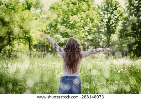 Cute little girl with the raised hands in air standing against the magic nature in sunny day, back to camera