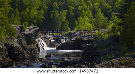 Water steps and falls down stone from a green forest into a blue river in Northern Minnesota.