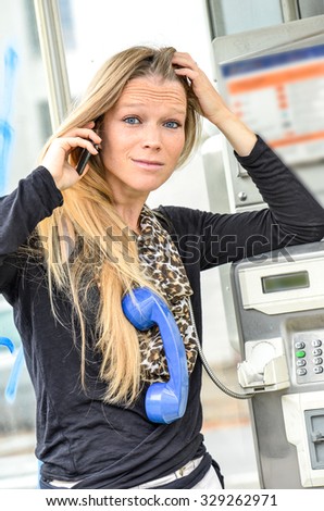 Beautiful fair hair woman talking with mobile holding blue phone around her neck in an old phone booth