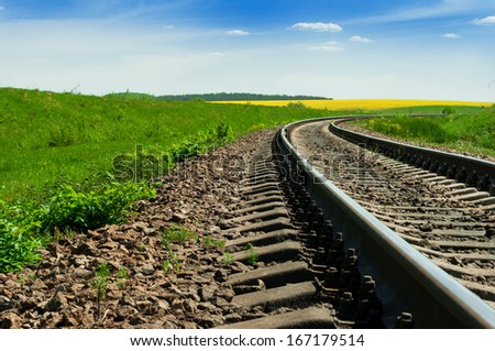 A railway turns upright through the spring green field. The sky is brightly blue with some white clouds