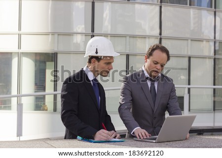 two architect working on computer wearing helmet glass building background